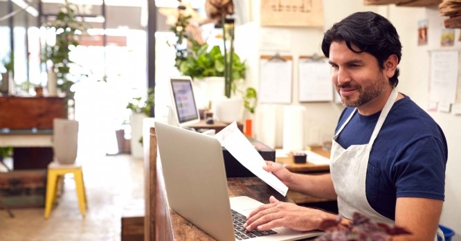 Male Sales Assistant Working On Laptop Behind Sales Desk Of Florists Store